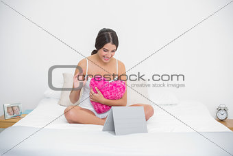 Charming young brown haired model in white pajamas hugging a pillow and looking at her tablet pc
