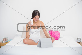 Happy young brown haired model in white pajamas holding a glass of milk while using tablet pc