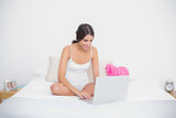 Concentrated young brown haired model in white pajamas using a laptop