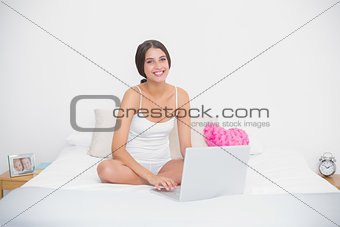 Charming young brown haired model in white pajamas using a laptop