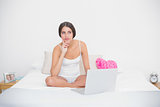 Thinking young brown haired model in white pajamas using a laptop
