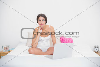 Thinking young brown haired model in white pajamas using a laptop