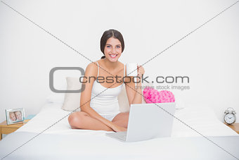 Cheerful young brown haired model in white pajamas drinking coffee while using laptop