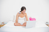 Joyful young brown haired model in white pajamas shopping online with her laptop