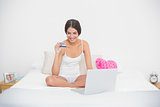 Smiling young brown haired model in white pajamas shopping online with her laptop