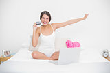 Cheering young brown haired model in white pajamas shopping online with her laptop