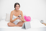 Concentrated young brown haired model in white pajamas texting with a mobile phone