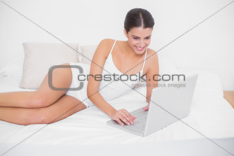 Pleased young brown haired model in white pajamas using a laptop