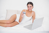 Happy young brown haired model in white pajamas shopping online with her laptop