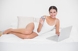 Relaxed young brown haired model in white pajamas shopping online with her laptop