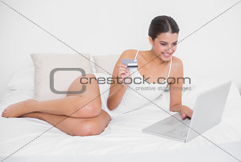 Pretty young brown haired model in white pajamas shopping online with her laptop