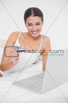 Cute young brown haired model in white pajamas shopping online with her laptop
