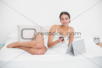 Thinking young brown haired model in white pajamas using a mobile phone