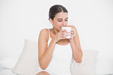 Calm young brown haired model in white pajamas enjoying coffee smell