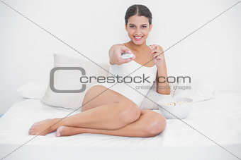 Cheerful young brown haired model in white pajamas watching tv while eating popcorn