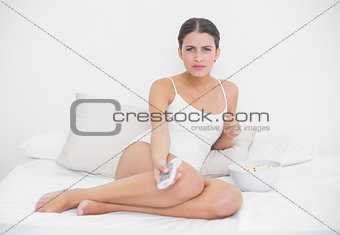 Frowning young brown haired model in white pajamas watching tv