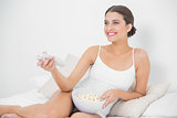 Attractive young brown haired model in white pajamas eating popcorn while watching tv