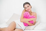 Seductive young brown haired model in white pajamas hugging a heart-shaped pillow