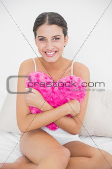 Romantic young brown haired model in white pajamas hugging a heart-shaped pillow