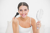 Happy young brown haired model in white pajamas applying powder on her face