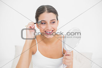 Pleased young brown haired model in white pajamas plucking her eyebrows