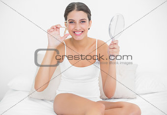 Charming young brown haired model in white pajamas using an eyelash curler