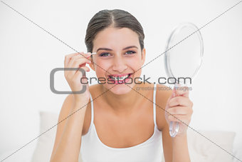 Amused young brown haired model in white pajamas plucking her eyebrows