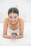 Amused young brown haired model in white pajamas looking at her mobile phone