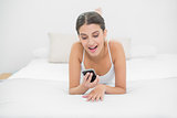 Natural young brown haired model in white pajamas looking at her mobile phone