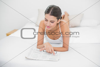 Attentive young brown haired model in white pajamas reading a newspaper