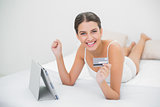 Successful young brown haired model in white pajamas shopping online with her tablet pc