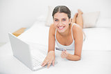 Charming young brown haired model in white pajamas shopping online with her laptop