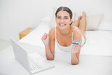 Victorious young brown haired model in white pajamas shopping online with her laptop