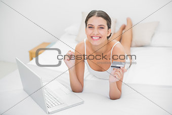 Victorious young brown haired model in white pajamas shopping online with her laptop