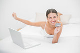 Excited young brown haired model in white pajamas shopping online with her laptop