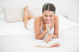 Calm young brown haired model in white pajamas reading a book
