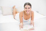 Delighted young brown haired model in white pajamas reading a book