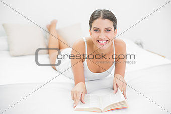 Delighted young brown haired model in white pajamas reading a book