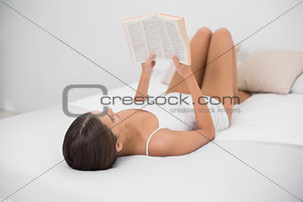 Peaceful young brown haired model in white pajamas reading a book