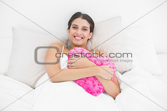 Content young brown haired model in white pajamas cuddling a heart shaped pillow