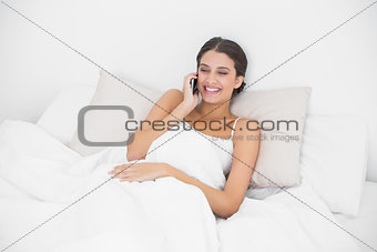 Laughing young brown haired model in white pajamas calling with her mobile phone