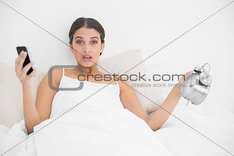 Astonished young brown haired model in white pajamas holding a mobile phone and an alarm clock