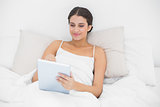 Cheerful young brown haired model in white pajamas using a tablet pc