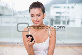 Pensive natural brown haired woman in white sportswear texting with her mobile phone