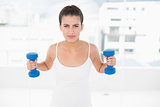 Frowning natural brown haired woman in white sportswear lifting dumbbells