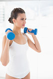 Thinking natural brown haired woman in white sportswear lifting dumbbells