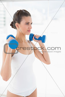 Thinking natural brown haired woman in white sportswear lifting dumbbells
