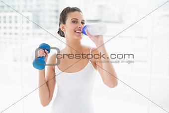 Cheerful natural brown haired woman in white sportswear drinking water while lifting a dumbbell