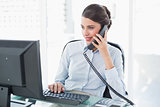 Concentrated classy brown haired businesswoman answering the telephone while using a computer