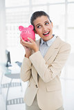 Surprised smart brown haired businesswoman shaking a piggy bank